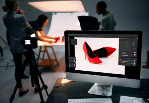 Photography team capturing product images of red shoes in a studio with a computer screen displaying the photo in editing mode